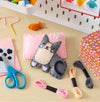 A Petit Collage Mini Craft Kit: Pocket Pet Kitten, including a handmade felt cat, pink pillow, embroidery floss, scissors, needle, orange felt fish, and colorful box, is displayed on a table with craft supplies in the background.
