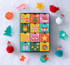 The Petit Collage 12 Crafts of Christmas Advent Calendar is displayed with felt ornaments—a tree, mitten, snowman, Santa, candy cane, star, cardinal, and pom-poms—on a snowy white background.