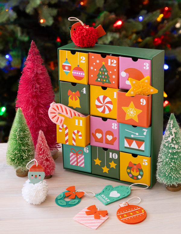 The 12 Crafts of Christmas Advent Calendar by Petit Collage sits on a table, its numbered drawers decorated and surrounded by festive ornaments, pink and green bottle brush trees, with a sparkling Christmas tree in the background.