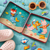 Two open Petit Collage Mix and Match Mermaids Magnetic Play Set tins showcase mermaid magnets arranged on an ocean backdrop, surrounded by toy pieces, pink slip-on shoes for kids, and flower-shaped sunglasses—all set on a teal quilted surface.