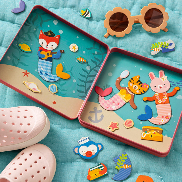 Two open Petit Collage Mix and Match Mermaids Magnetic Play Set tins showcase mermaid magnets arranged on an ocean backdrop, surrounded by toy pieces, pink slip-on shoes for kids, and flower-shaped sunglasses—all set on a teal quilted surface.