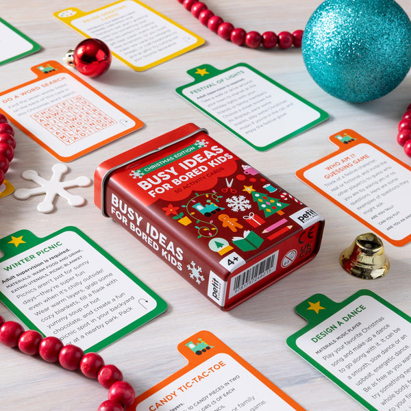 A deck of Petit Collage’s Busy Ideas for Bored Kids: Christmas Edition rests on a table, surrounded by Christmas ornaments and assorted holiday-themed activity cards for children.