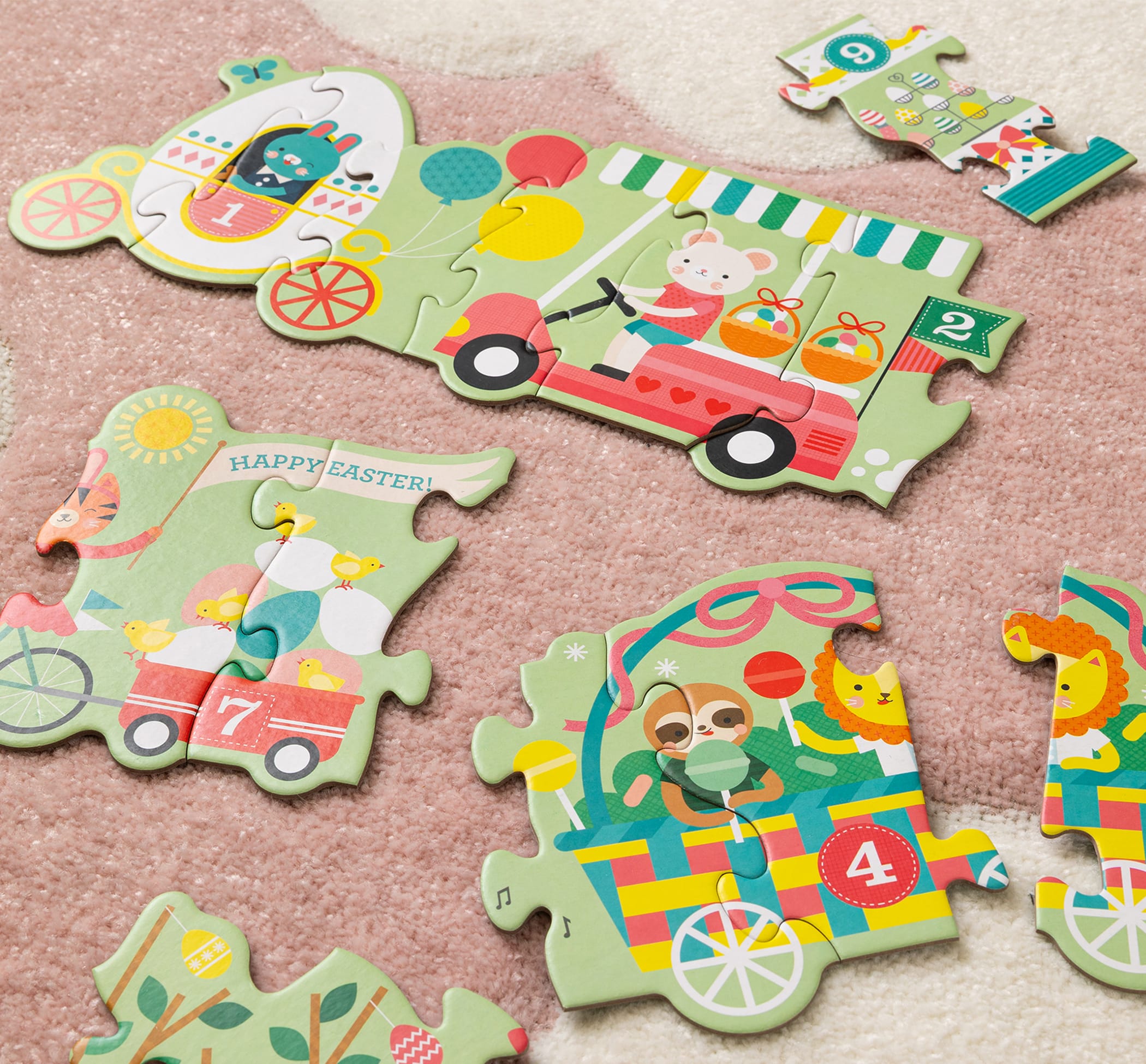 Pieces from the Petit Collage Easter Parade Shaped Floor Puzzle, with colorful cartoon animals and numbered vehicles, are scattered on a pink carpet.