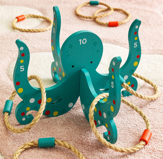 The Petit Collage Happy Octopus Wooden Hoopla, a wooden ring toss game with numbered arms and rope rings, is displayed on a pink and white rug.