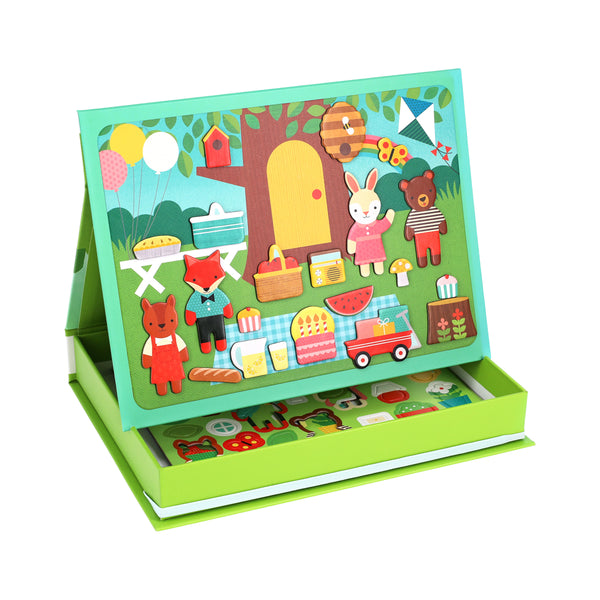 The Petit Collage Treehouse Party Magnetic Play Scene includes animal characters, picnic items, and outdoor scenery that attach to an upright board, plus extra magnetic pieces stored in the box below.