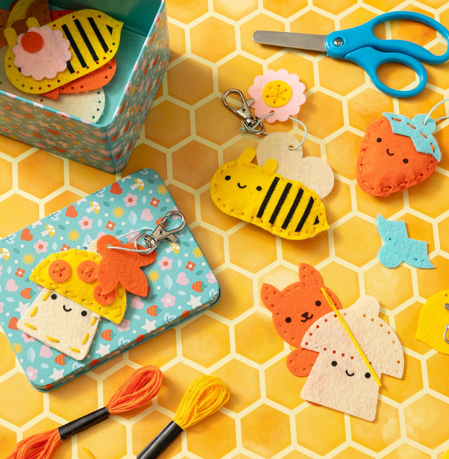 Brightly colored felt keychains shaped like a bee, cat, strawberry, and taco from the Petit Collage Mini Craft Kit: Pocket Pal Bag Charms, shown with included craft supplies and a patterned tin on a honeycomb-patterned surface.