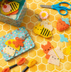 Brightly colored felt keychains shaped like a bee, cat, strawberry, and taco from the Petit Collage Mini Craft Kit: Pocket Pal Bag Charms, shown with included craft supplies and a patterned tin on a honeycomb-patterned surface.