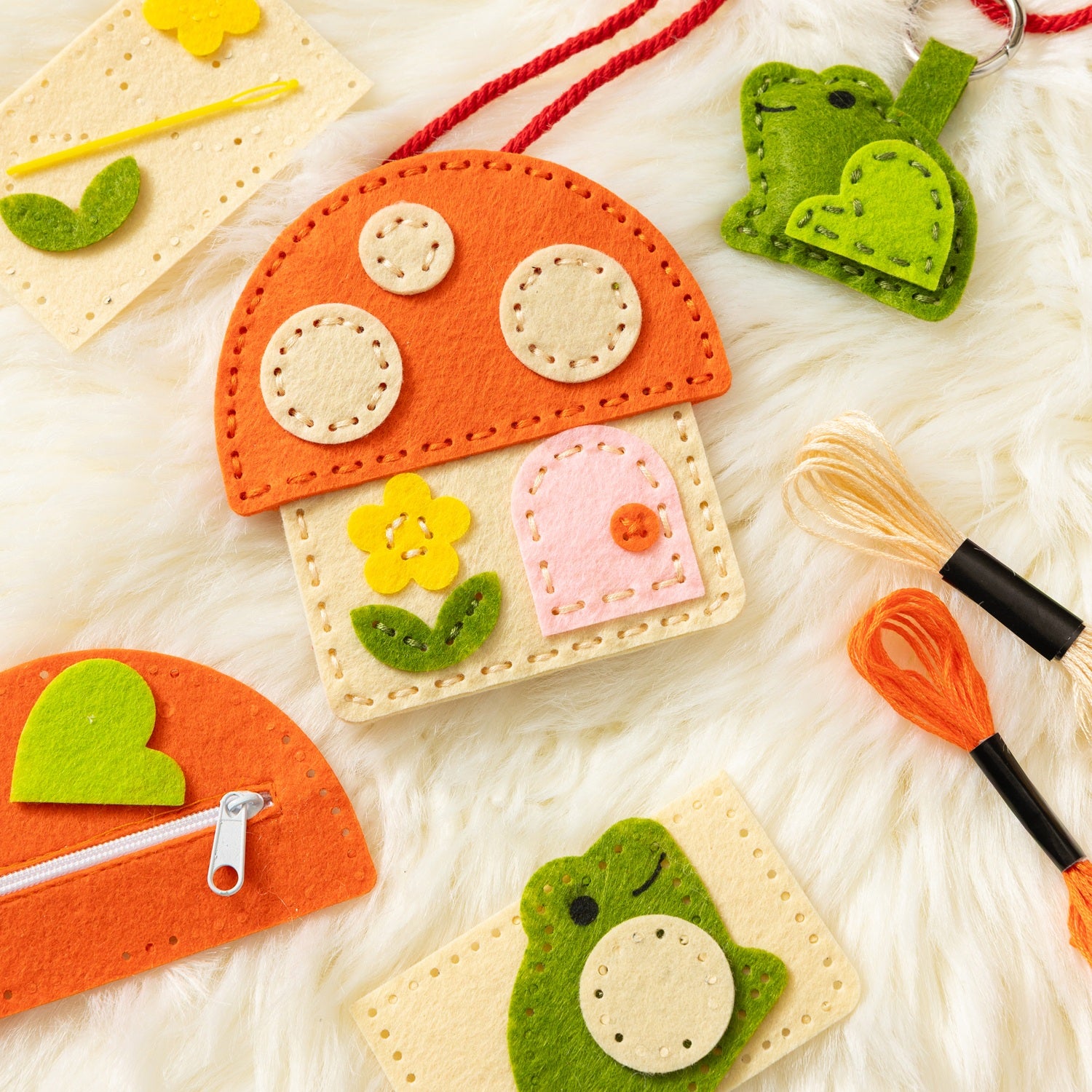 A variety of felt craft accessories from Petit Collage are displayed on a soft white background, featuring the Mini Craft Kit: Toadstool Purse, along with a green frog keychain, a small zippered pouch, thread bundles, and stitched felt pieces.