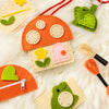 A variety of felt craft accessories from Petit Collage are displayed on a soft white background, featuring the Mini Craft Kit: Toadstool Purse, along with a green frog keychain, a small zippered pouch, thread bundles, and stitched felt pieces.