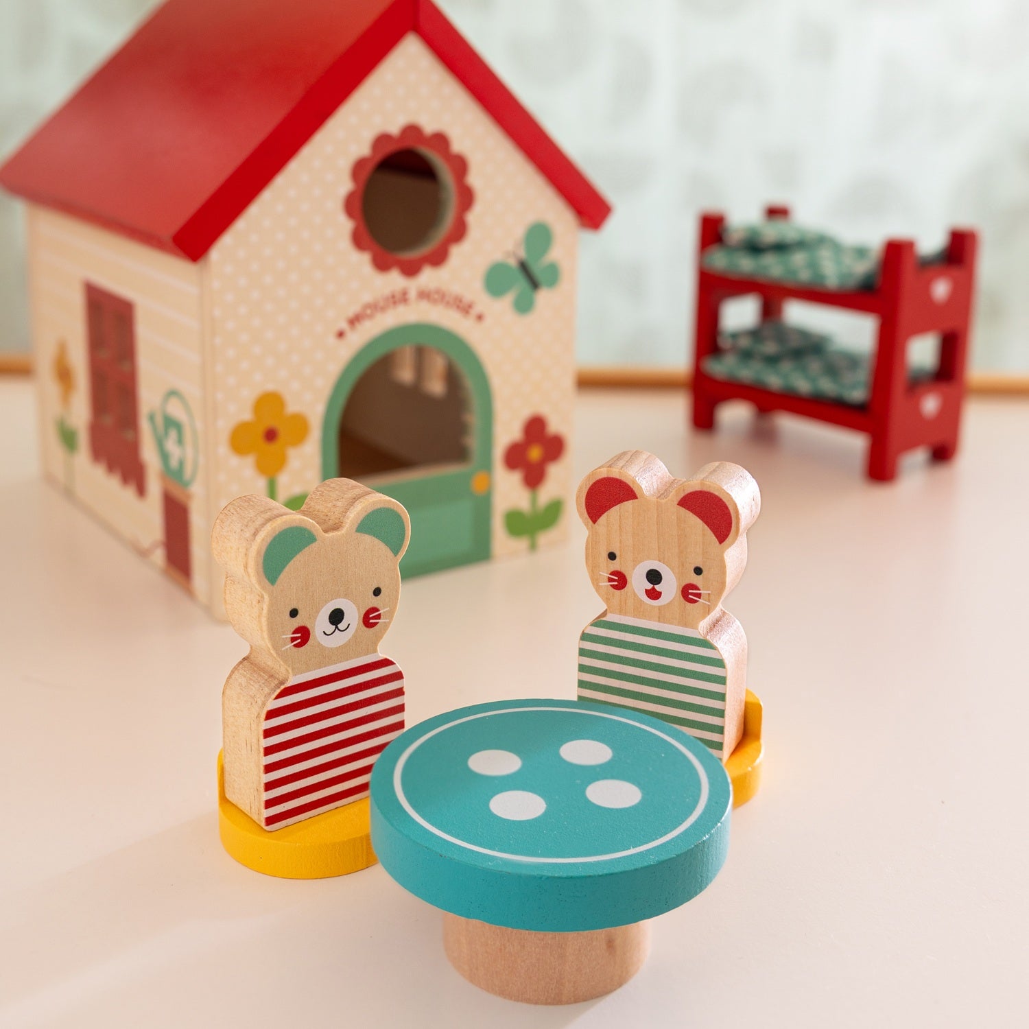 Two wooden mice from the Petit Collage Mouse House Wooden Play Set sit at a small round table in front of their toy house, with a red shelf holding small blocks in the background.