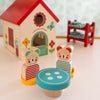 Two wooden mice from the Petit Collage Mouse House Wooden Play Set sit at a small round table in front of their toy house, with a red shelf holding small blocks in the background.