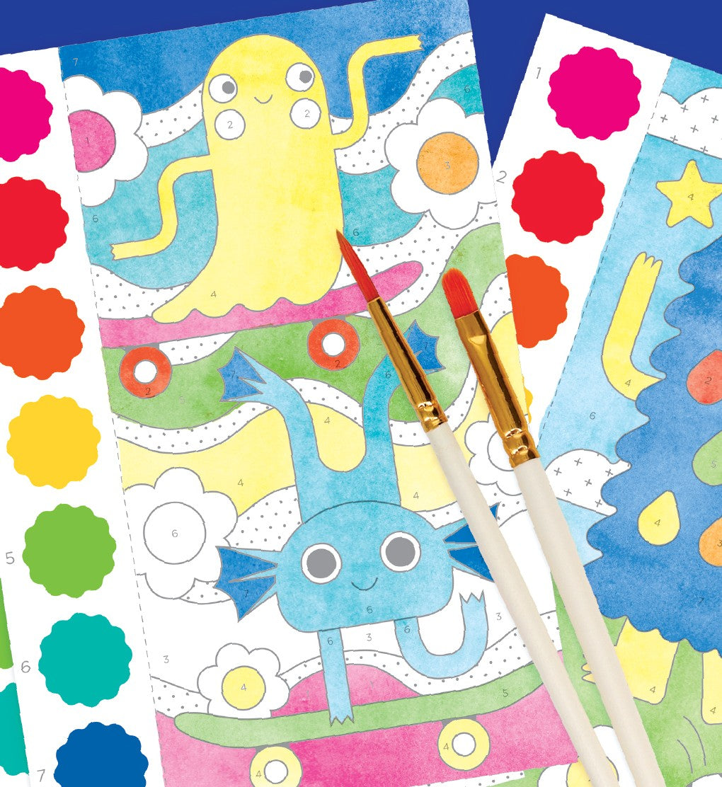 Two paintbrushes rest on colorful Petit Collage Paint by Numbers: Monsters sheets, featuring cartoon creatures, with color guide stickers visible beside the illustrations.