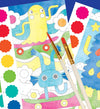 Two paintbrushes rest on colorful Petit Collage Paint by Numbers: Monsters sheets, featuring cartoon creatures, with color guide stickers visible beside the illustrations.