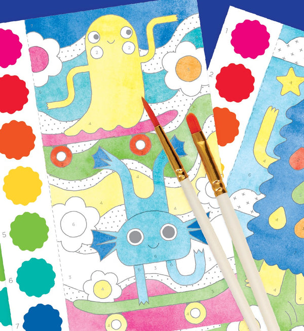 Two paintbrushes rest on colorful Petit Collage Paint by Numbers: Monsters sheets, featuring cartoon creatures, with color guide stickers visible beside the illustrations.