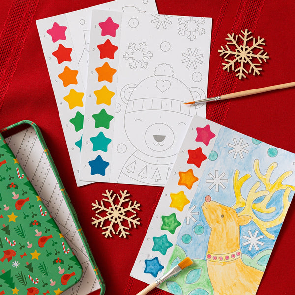 Petit Collage’s Paint by Numbers: Christmas set features watercolors, unfinished and completed holiday animal paintings, paintbrushes, and decorative snowflakes arranged on a red tablecloth.
