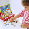A child in a red and white striped shirt arranges animal pieces from the Petit Collage Pet Hospital Magnetic Play Scene on a table next to its colorful illustrated board.
