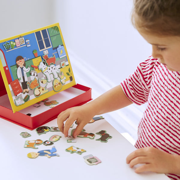 A child in a red and white striped shirt arranges animal pieces from the Petit Collage Pet Hospital Magnetic Play Scene on a table next to its colorful illustrated board.