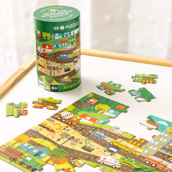 A partially assembled Spot the Difference Puzzle: Roads and Rails by Petit Collage sits on a table, with its green cylindrical box beside it. The 49-piece puzzle is designed for ages 4 and up.