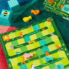 A vibrant board game called Slides and Climbs: A Get to the Top Game by Petit Collage features animal illustrations, wooden bird and fish tokens, a green box displaying the game’s name, and two patterned blocks arranged on a multicolored carpet.