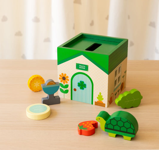 The Petit Collage Petit Pals: Tilly Tortoise set features a green and white toy house-shaped box on a table, surrounded by colorful wooden pieces like a turtle, leaf, and flower.