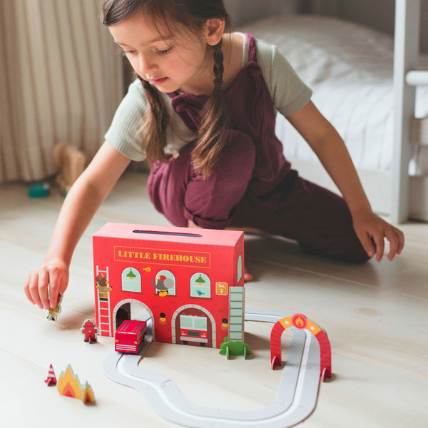 A young child plays on the floor with the Petit Collage Wind Up & Go Fire Station Play Set, moving small figures and a fire truck along the track.